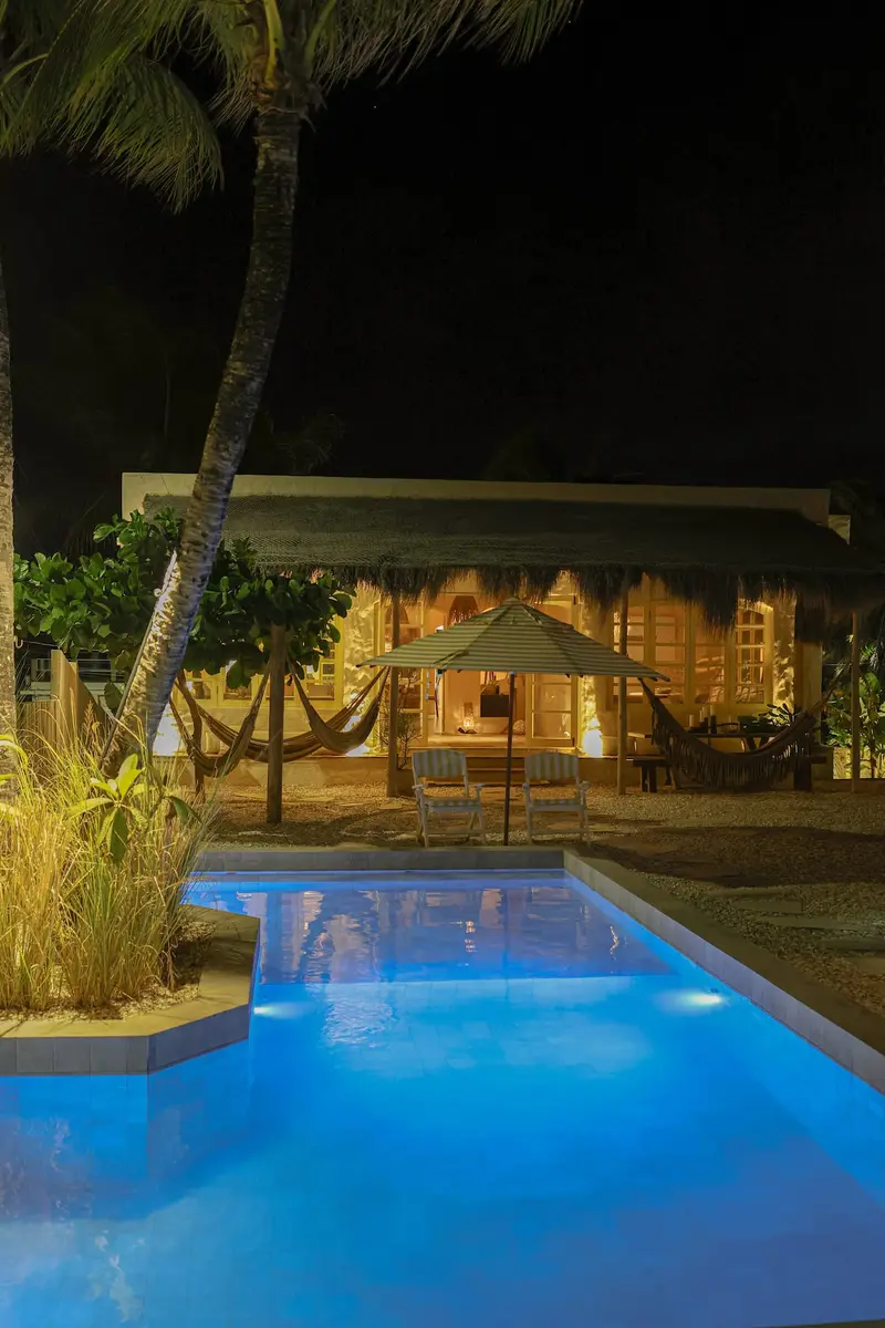 Piscina azul acesa à noite, com a casa iluminada ao fundo