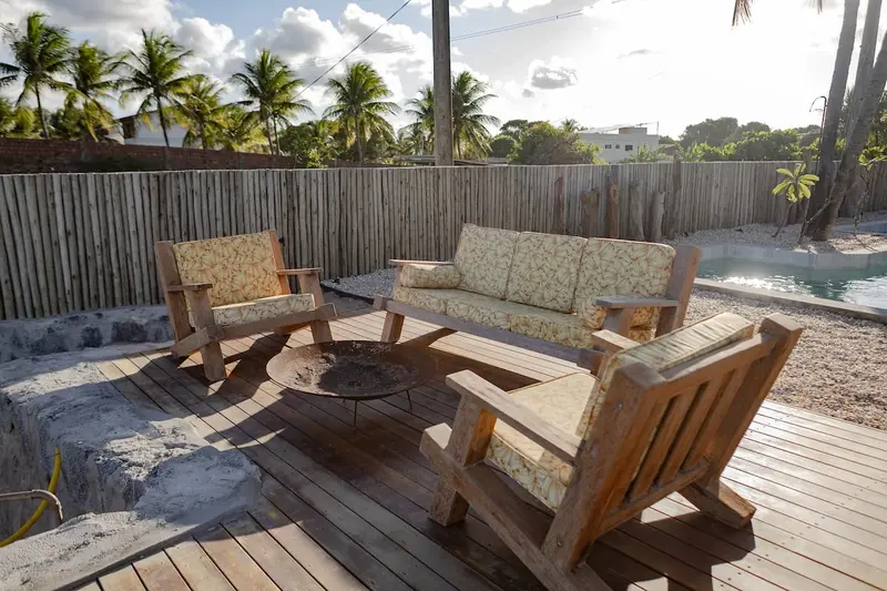 Poltronas e sofá de madeira ao redor do fire pit, com coqueiros ao fundo