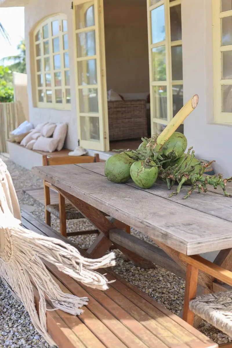 Mesa comprida de madeira na varanda, com cocos verdes e redes ao lado